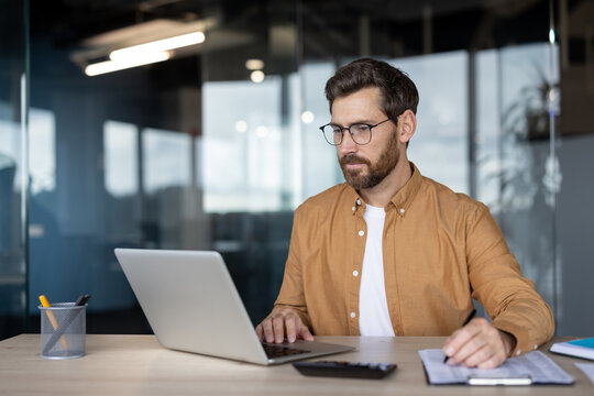 Fototapeta Businessman in glasses and a brown shirt is actively working at his office desk, typing on a laptop and writing notes, demonstrating focus on work, productivity, and professional multitasking
