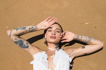 Portrait of a beautiful young woman dressed in white lying on the beach on the wet sand sunbathing. The waves break on her wet body and she cools off on a sunny summer day. Photo taken from above.