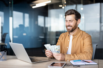 Bearded male entrepreneur smiling, holding and counting cash while working at a laptop on a desk in a modern office, symbolizing financial success, profit, and payment