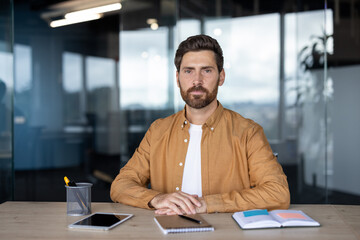 Bearded male entrepreneur in a modern office, sitting at desk with tablet and notebook, looking...