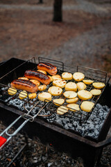Savory sausages and sliced potatoes are grilling on a wire rack over hot charcoal embers in an outdoor barbecue grill. This image captures the rustic experience of cooking outdoors 