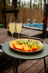 Two champagne flutes and a plate of sliced peaches and grapes are served on a small table on a wooden deck, with a hot tub and a bamboo fence blurred in the background, signaling relaxation.