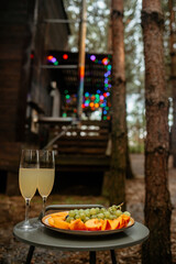 Two champagne flutes and a plate of sliced peaches and grapes sit on an outdoor table, set against a cozy wooden cabin decorated with colorful string lights in a rustic forest setting.