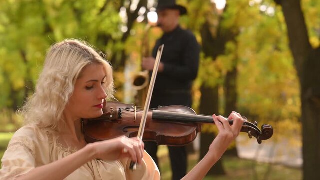 A beautiful blonde violinist and a saxophonist perform melodies in an autumn park. A fantastic romantic mood, a touch of autumn. Musical instruments playing at an outdoor concert. A musical duet.	
