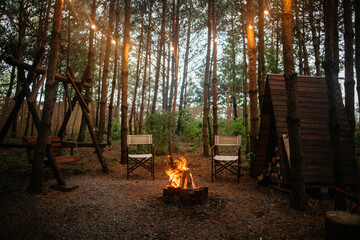 A warm, inviting campsite scene at dusk with a blazing campfire, two empty chairs, a rustic wooden swing, and a firewood shed, all illuminated by string lights hanging in the pine trees.
