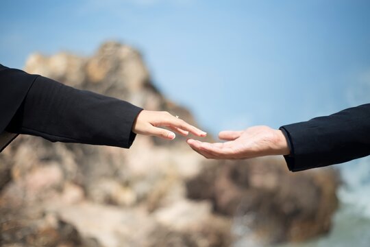 Two hands, reaching, one with a ring, against a blurred rocky landscape under a bright blue sky, captured in a soft, hopeful, cinematic style.
