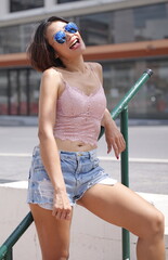 Asian woman leans casually on a green metal railing, smiling in pink lace crop top, denim shorts, and reflective sunglasses—stylish, relaxed, and framed by a casual urban street setting