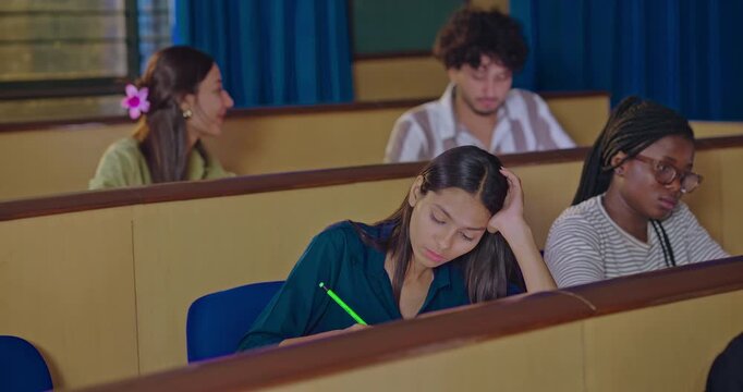 Indian young adult gen z dull pupil girl hold pencil sitting writing do study exam notes indoor class room. Lady female bad tired lazy sleepy lack energy need rest sleep on desk table college place