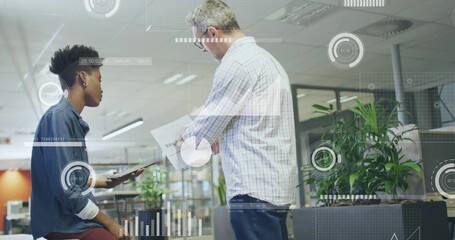 Reviewing tablet and papers, two colleagues meeting in open-plan office, with data overlays