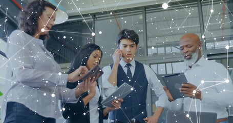 Collaborating four business professionals standing in glass-walled office, using networked tablets