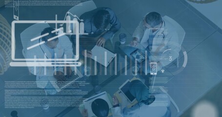 Collaborating team leaning over charts around glass table in office, with laptops and stethoscopes