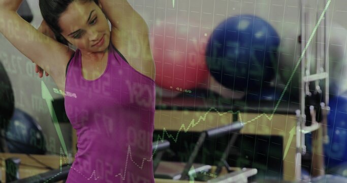 Stretching woman in pink tank top reaching overhead in gym, with exercise balls and market graphs