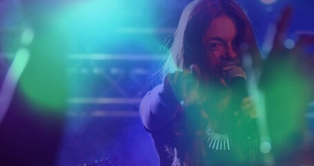 Performing singer wearing necklace holding microphone on stage under spotlights, with blurred hands