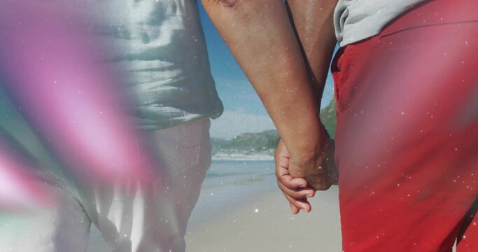 Walking couple wearing swimwear, holding hands and strolling beach shoreline, with pink lens flare
