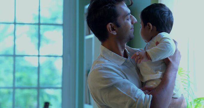 Indian trying to make baby infant boy sleep while gently rocking him in arms near breezy airy window inside home during quiet afternoon moment of parental affection and bonding time