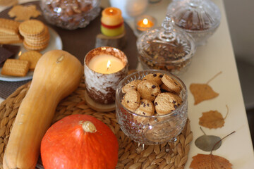 Lit candles, cookies, chocolate, nuts, tea, wine, pumpkins, books, reading glasses and autumn leaves on the table. Autumnal hygge at home. Selective focus.