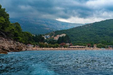 Fototapeta premium Panorama of resort town against backdrop of dramatic sea and cloudy sky, Petrovac, Montenegro, beautiful view during a boat trip, view from the sea