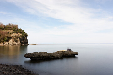 Rocky Outcrops Against a Calm Sea. Rock formations extending into calm, glassy waters near a cliff with green vegetation. The soft light and cloudy sky enhance the peaceful coastal landscape.