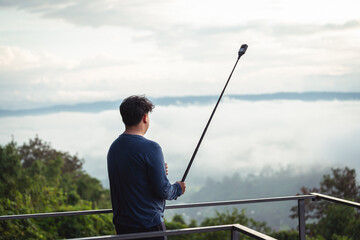 Young Man Using Selfie Stick to Capture Scenic View from Observation Deck Surrounded by Clouds and Lush Green Trees on a Beautiful Day