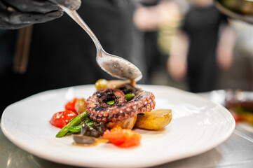Chef garnishing gourmet octopus dish with roasted potatoes, green beans, and cherry tomatoes in a...