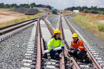 Two railway engineers in safety helmets and reflective jackets inspect rail alignment and fasteners on a new track. They discuss project details using blueprints, emphasizing teamwork and precision.