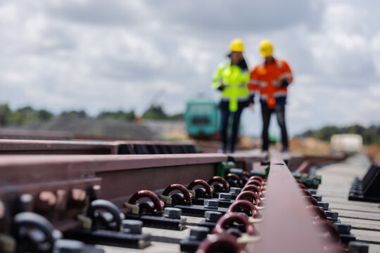 Close-up of railway bolts and clips securing steel rails on concrete sleepers, with two engineers in safety gear inspecting in the background. 