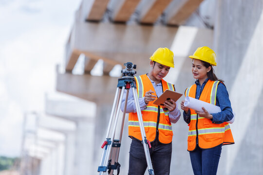 Two civil engineers wearing helmets and reflective vests inspect bridge construction plans using a tablet and blueprints beside a surveying tripod. 