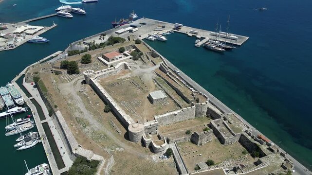 Aerial view of the imposing Neratzia Castle, its stone walls contrasting with the turquoise sea and docked boats, Kos, Greece.