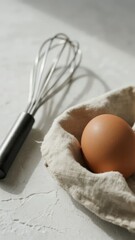 A single brown egg rests in a cloth-lined nest beside a metal whisk on a textured white surface.