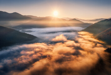 Aerial view of mountains in low clouds at sunrise in autumn. Mountain valley in fog in fall at dawn. Beautiful landscape with hills, forest, sun, colorful sky. Top drone view of foggy hills