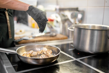 Chef seasoning mushrooms in stainless steel pan on electric stove in professional kitchen. Culinary preparation with fresh ingredients and cooking utensils in background.