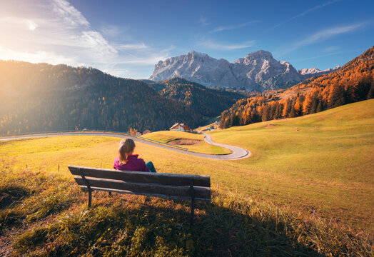 Young woman sitting on the bench and beautiful alpine village at sunset in autumn. Dolomites, Italy. Colorful landscape with girl, meadows and hills, orange trees, road, mountain, blue sky in fall