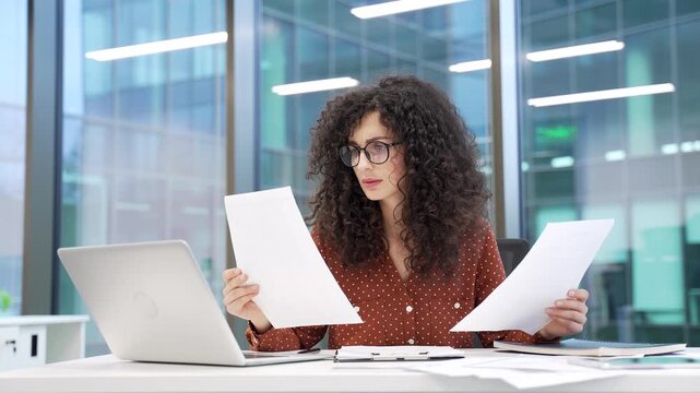 Frustrated businesswoman having difficulty with paperwork while sitting at workplace in modern business office. Puzzled disappointed female employee looks at documents and cannot understand problem