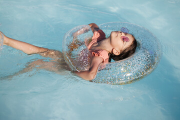 Cute funny toddler girl in a sunglasses relaxing on inflatable toy ring floating in pool have fun during summer vacation. Child having fun in swimming pool. 