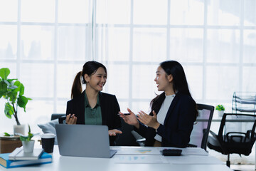 Two smiling businesswomen engaging in a lively discussion while working together at a desk with a laptop and documents, emphasizing teamwork and communication in a professional setting