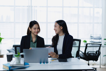 Two smiling Asian businesswomen engaging in a productive conversation, collaborating on a project, and discussing work while looking at a tablet in a bright modern office