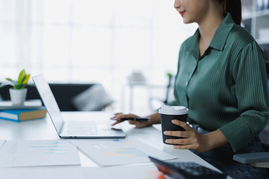 Young professional woman at her office desk, working on a laptop while holding a takeaway coffee cup, focused on remote work tasks, typing, planning, and research