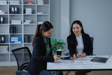 Two businesswomen collaborate at a desk in a modern office, reviewing financial reports, laptop and calculator nearby, discussing strategy and analyzing charts for growth and planning