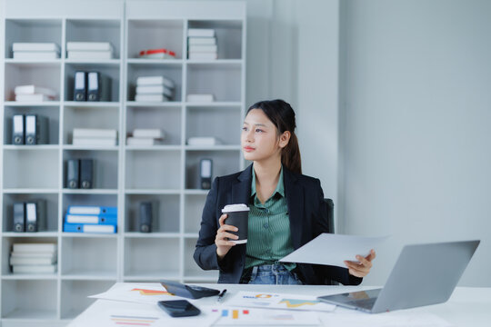 Asian businesswoman holding a coffee cup and documents while looking away, sitting at a desk with a laptop and office supplies, working and planning in a modern office environment