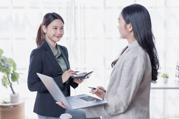 Two Asian businesswomen collaborate in an office, analyzing financial data with a laptop and calculator, discussing charts and paperwork while planning strategy and growth