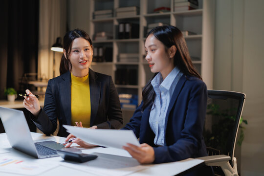 Asian business women collaborating on a project in a modern office, discussing documents and points on a laptop screen while working overtime at night