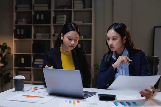 Two young Asian businesswomen are collaborating. Reviewing documents and using a laptop together at their desk. Working overtime in a modern office environment late at night. Focusing on project data