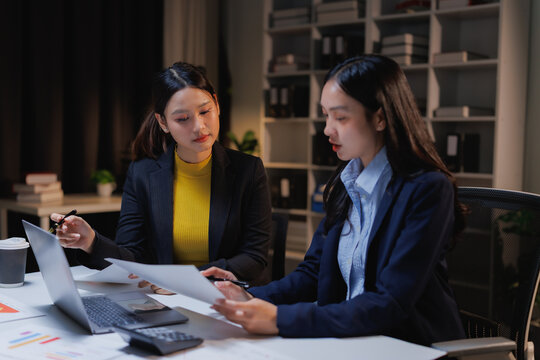 Two young Asian businesswomen are collaborating on a project, examining financial documents and working on a laptop at their office desk, focusing on the task during late hours