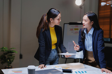 Asian businesswomen collaborating, analyzing charts and documents on a desk, actively engaging in discussion and brainstorming during a professional business meeting in an office setting