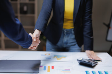 Business partners shaking hands over a table with financial charts and a laptop, signifying a successful agreement, partnership, and collaboration in the office