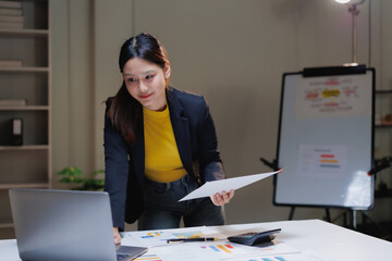 Asian businesswoman standing at a desk, reviewing charts and graphs on documents and a laptop, discussing financial data during a remote business meeting in a professional office