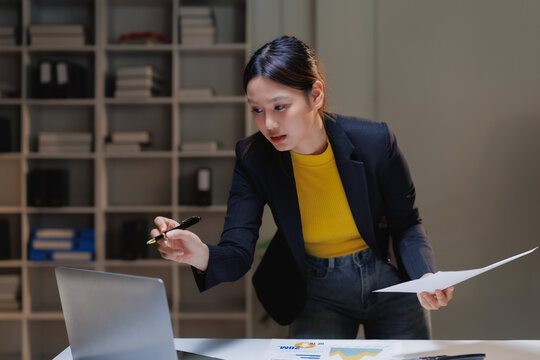 Asian businesswoman concentrating on an important project, pointing at a laptop screen while reviewing documents and working diligently in the office during evening hours - Powered by Adobe