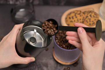 Close up. A woman's hands pours coffee beans into a coffee grinder from a transparent glass with a black spoon. Oatmeal cookies with chocolate chips on a wooden board. Dark background