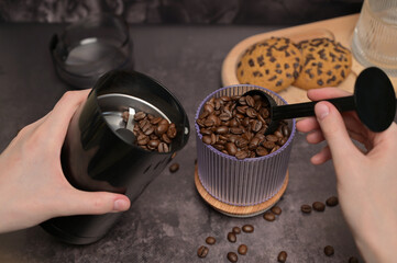 Close up. A woman's hands pours coffee beans into a coffee grinder from a transparent glass with a black spoon. Oatmeal cookies with chocolate chips on a wooden board. Dark background