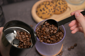Close up. A woman's hand pours coffee beans into a coffee grinder from a transparent glass with a black spoon. Oatmeal cookies with chocolate chips on a wooden board. Dark background